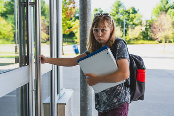 Sad/depressed teen girl/student opening a glass door to her school while wearing a backpack and holding binders.