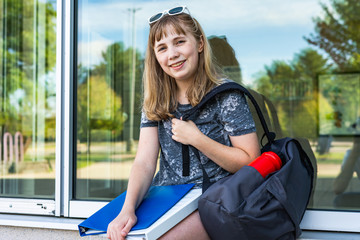 Happy/smiling teen girl/student sitting on a window ledge of her school while holding binders and...