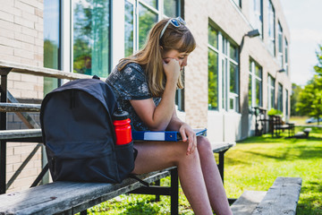 Depressed/Sad teen girl sitting on a set of bleachers next to her school with a backpack and binders.