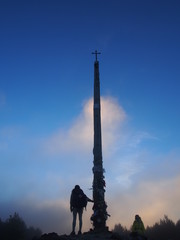 Fototapeta premium Pilgrim and beautiful blue sky with Cruz de Ferro (Cross of Iron) in the early morning, Camino de Santiago, Way of St. James, Foncebadon to Ponferrada, French way, Spain