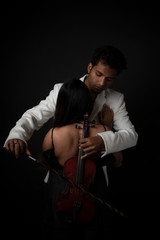 Fototapeta na wymiar An young and attractive Indian brunette Bengali man in white western suit playing violin resting on his partner's bare back in black studio copy space background. Indian fashion portrait and lifestyle