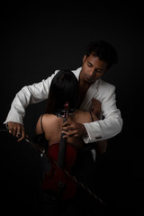 Obraz na płótnie Canvas An young and attractive Indian brunette Bengali man in white western suit playing violin resting on his partner's bare back in black studio copy space background. Indian fashion portrait and lifestyle