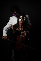 Fototapeta na wymiar Young and attractive Indian brunette Bengali man with mask in western suit playing violin resting on his partner's body in black studio copy space background. Indian fashion portrait and lifestyle