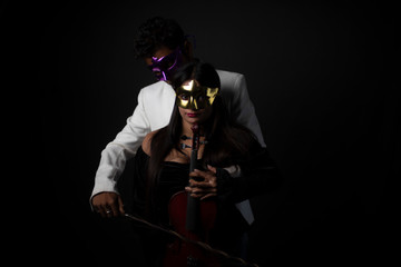 Young and attractive Indian brunette Bengali man with mask in western suit playing violin resting on his partner's body in black studio copy space background. Indian fashion portrait and lifestyle