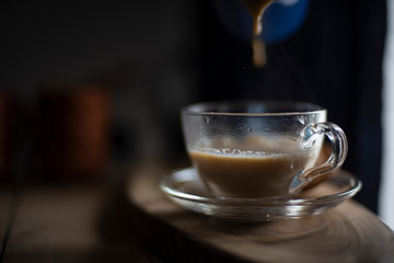 Hot infused tea/coffee being poured in a transparent glass cup and soccer kept in a wooden tray/table from a kettle in black background in the winter morning. Indian beverages and food photography.