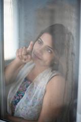 Blurred portrait of an young Indian Bengali brunette girl in white sleeping wear standing in front of a wet glass window with dewdrops on it in a winter morning. Indian lifestyle.