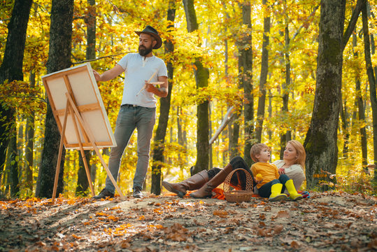Handsome bearded man draws a picture of his young family in the park using a palette with paints and a spatula. Drawing easel and canvas with a picture of closely related people.