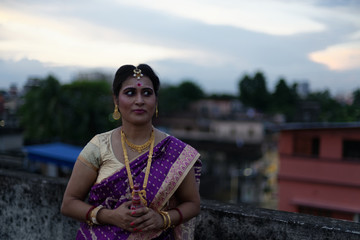 Fototapeta na wymiar Portrait of a beautiful smiling brunette Indian Bengali bride in traditional sari standing on the roof top in afternoon under blue sky. Indian lifestyle.