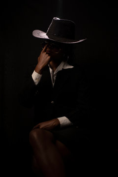 An young and attractive dark skinned Indian Bengali brunette woman in western jacket, hat removing goggles while sitting under spotlight in a black copy space background. Indian fashion photography.
