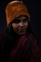 Portrait of an young and attractive dark skinned Indian woman wearing woolen cap and jacket feeling cold in a  winter night in dark background. Winter and Christmas