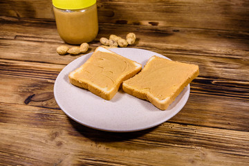 Glass jar with peanut butter and plate with sandwiches on a wooden table