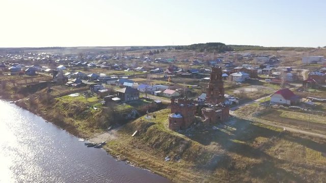 Aerial view of old ruined church located at the river shore near the small village. Clip. Abandoned old ruins of the chapel and houses on blue cloudy sky background.