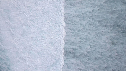 Above the ocean, aerial lock down view at foaming waves and surface near coast line