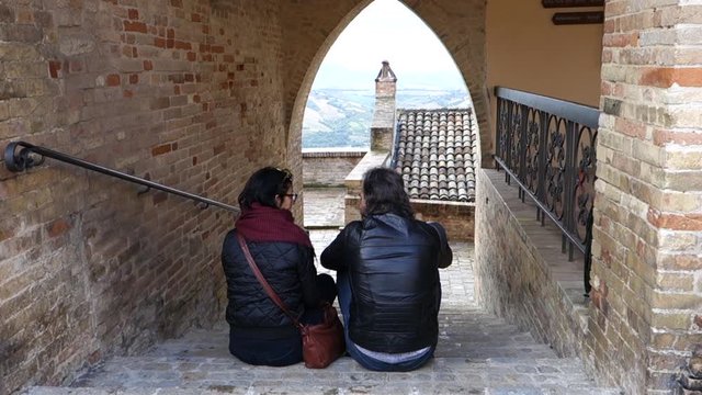 A loving couple looking at the phone sitting on the steps of an ancient Italian village in front of the arch with a panoramic view. Close up, back view, togetherness concept
