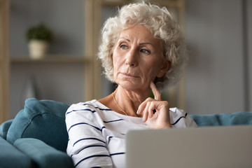 Pensive aged woman sitting on sofa with laptop look away