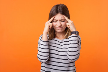Portrait of stressed young woman with brown hair in long sleeve striped shirt standing with closed eyes holding temples, frowning suffering headache. indoor studio shot isolated on orange background