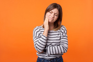 Portrait of unhappy sick young woman with brown hair in long sleeve striped shirt standing, touching cheek, suffering acute toothache, need dentist indoor studio shot isolated on orange background