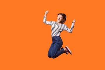 Fotobehang Full length portrait of excited pretty woman with brown hair in casual shirt and denim jumping celebrating victory, raising fists showing yes gesture. indoor studio shot isolated on orange background © khosrork