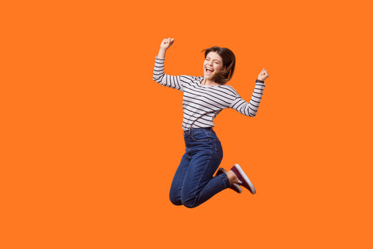 Full length portrait of excited pretty woman with brown hair in casual shirt and denim jumping celebrating victory, raising fists showing yes gesture. indoor studio shot isolated on orange background