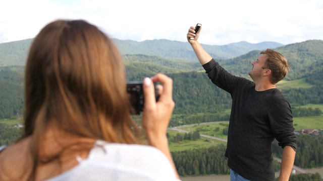 Tourists couple enjoying the nature in mountains, young woman taking photos of man that takes selfie on the phone.