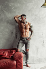 Fashion portrait of Sexy naked male model with tattoo and a black beard standing in hot pose on red leather sofa. Loft room interior with grey concrete wall. Professional Studio image.