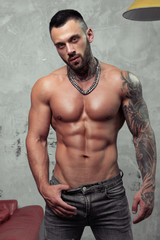 Fashion portrait of Sexy naked male model with tattoo and a black beard standing in hot pose. Loft room interior with grey concrete wall. Professional Studio image.