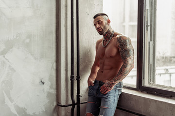 Fashion portrait of Sexy naked male model with tattoo and a black beard standing in hot pose on near the window. Loft room interior with grey concrete wall. Professional Studio image.