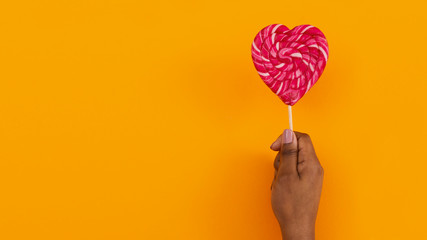 Black female hand holding heart shaped lollipop on orange background