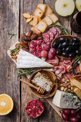 Traditional italian antipasto plate. Assorted cheeses on wooden cutting board. Brie cheese, cheddar slices, gogonzola, walnuts grapes, olives, prosciutto, rosemary and glass of red wine. top view