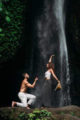 A man proposes to a woman at a waterfall. A guy proposes to a girl in Bali. Offer of a hand in travel. Honeymoon trip. Declaration of love. A couple in love on a waterfall. Happy couple in Bali