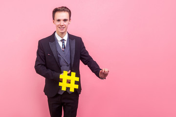 Portrait of elegant happy man in suit with stylish haircut holding large yellow hashtag symbol and directing another hand at empty copy space, place for your text, list of hashtags. indoor studio shot