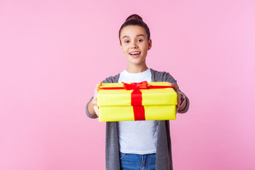 Share holiday present. Portrait of generous teenage brunette girl with bun hairstyle in casual clothes giving gift box to camera and smiling, charity donation. studio shot isolated on pink background