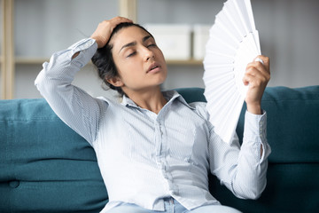 Tired indian woman using fan sit on couch at home
