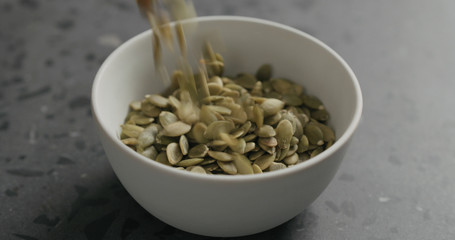 closeup dried pumpkin seeds falling into white bowl on terrazzo countertop