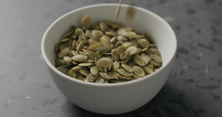 closeup dried pumpkin seeds falling into white bowl on terrazzo countertop