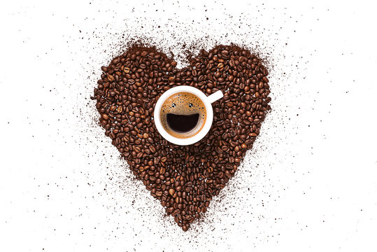 heart made from coffee beans and ground coffee on a white background. in the center is a cup, in a cup from coffee foam a happy smiling face. invigorating drink concept