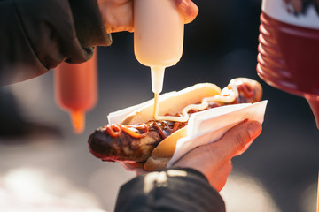Woman pouring tomato sauce, mayonnaise and mustard on a grilled pork sausage Bratwurst at a German Market. - obrazy, fototapety, plakaty