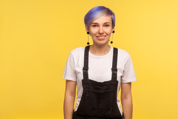 Portrait of happy trendy hipster woman with violet short hair in overalls looking at camera with toothy smile, positive mood, stylish fashionable appearance. isolated on yellow background, studio shot