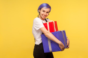 Portrait of glad positive hipster girl with violet short hair in overalls carrying gift boxes and smiling at camera, embracing holiday presents. isolated on yellow background, indoor studio shot