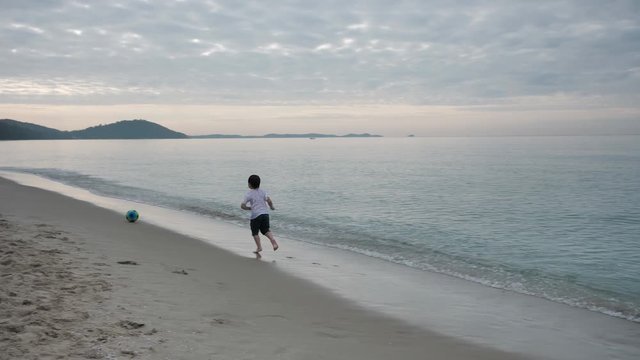 Happy Asian children boy playing football On Summer Vacation on sand in the beach In the morning time, sunrise. Holiday and Travel concept. Slow motion