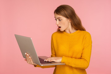 Fototapeta na wymiar Portrait of astonished ginger girl in sweater holding laptop and working, typing on keyboard and looking with amazement and shock, using computer. indoor studio shot isolated on pink background