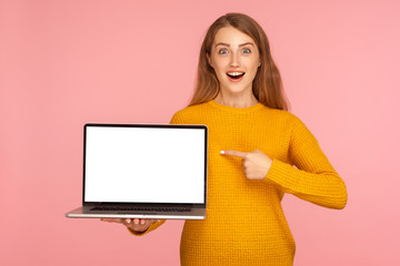 Shocking advertising! Portrait of amazed ginger girl in sweater pointing at laptop empty screen and...