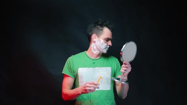 A man with foam on his face looks at the camera and begins to shave with a razor