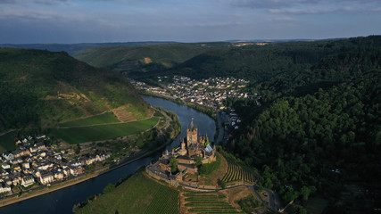 Fototapeta na wymiar Aerial view of Cochem Castle on the hill near river surrounded by green vineyards, trees, fields, small houses, bridge. German flag waving on the top of tower. Summer. Germany historical landmarks.