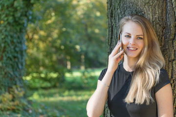 Smiling attractive young girl is phoning in the park. All potential trademarks are removed.