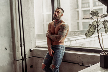 Fashion portrait of Sexy naked male model with tattoo and a black beard standing in hot pose on near the window. Loft room interior with grey concrete wall. Professional Studio image.