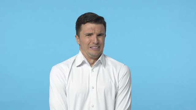 Slow-motion adult caucasian man in white shirt cringing, grimacing from aversion, feeling strong disgust, shivering from bad feeling, standing reluctant and disgusted, want puke, blue background