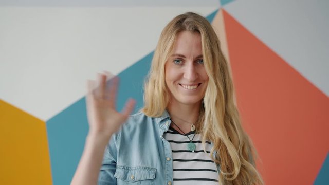 Portrait of cute blond girl waving hand smiling in friendly way on colorful background greeting welcoming people. Gestures and lovely people concept.