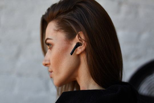 Young brown hair street fashion woman in black hoodie with opened neck turned back and uses to talk or listen music wireless earphones in ear in loft interior with white brick wall