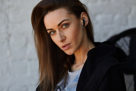 Young depressed street fashion woman in black hoodie and grey print shirt use to talk or listen music wireless earphones in ear in loft interior and fixes brown hair and looks to camera